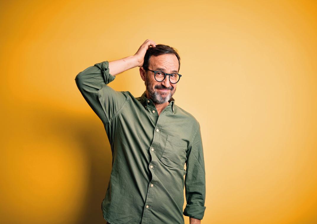 Deze afbeelding toont een man met een bril en een groene overhemd staat voor een felgele achtergrond.