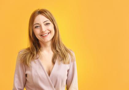 Vrouw met lang, lichtbruin haar glimlacht in een lichte blouse tegen een gele achtergrond.